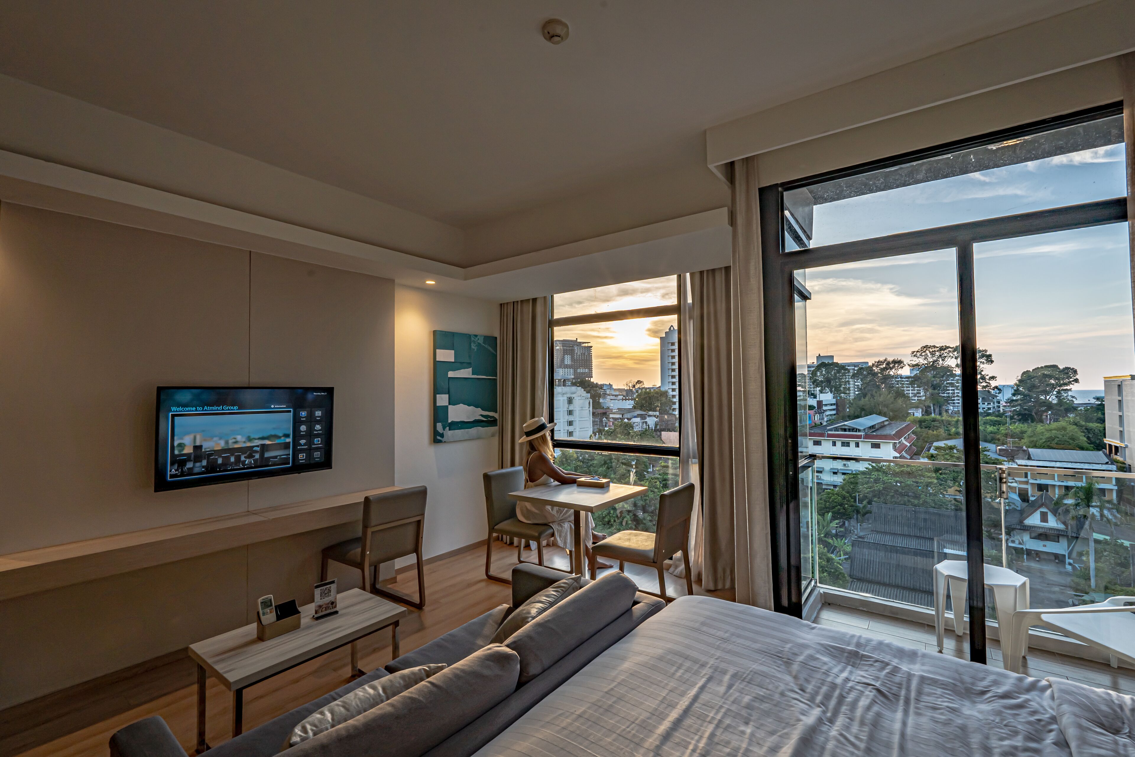 A modern hotel room with a large bed, a desk, a chair, a TV mounted on the wall, and a large window offering a city view.