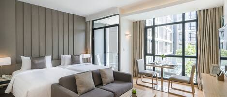 A modern hotel room with a large bed, a sofa, a small table, and a dining area with a view of the city.