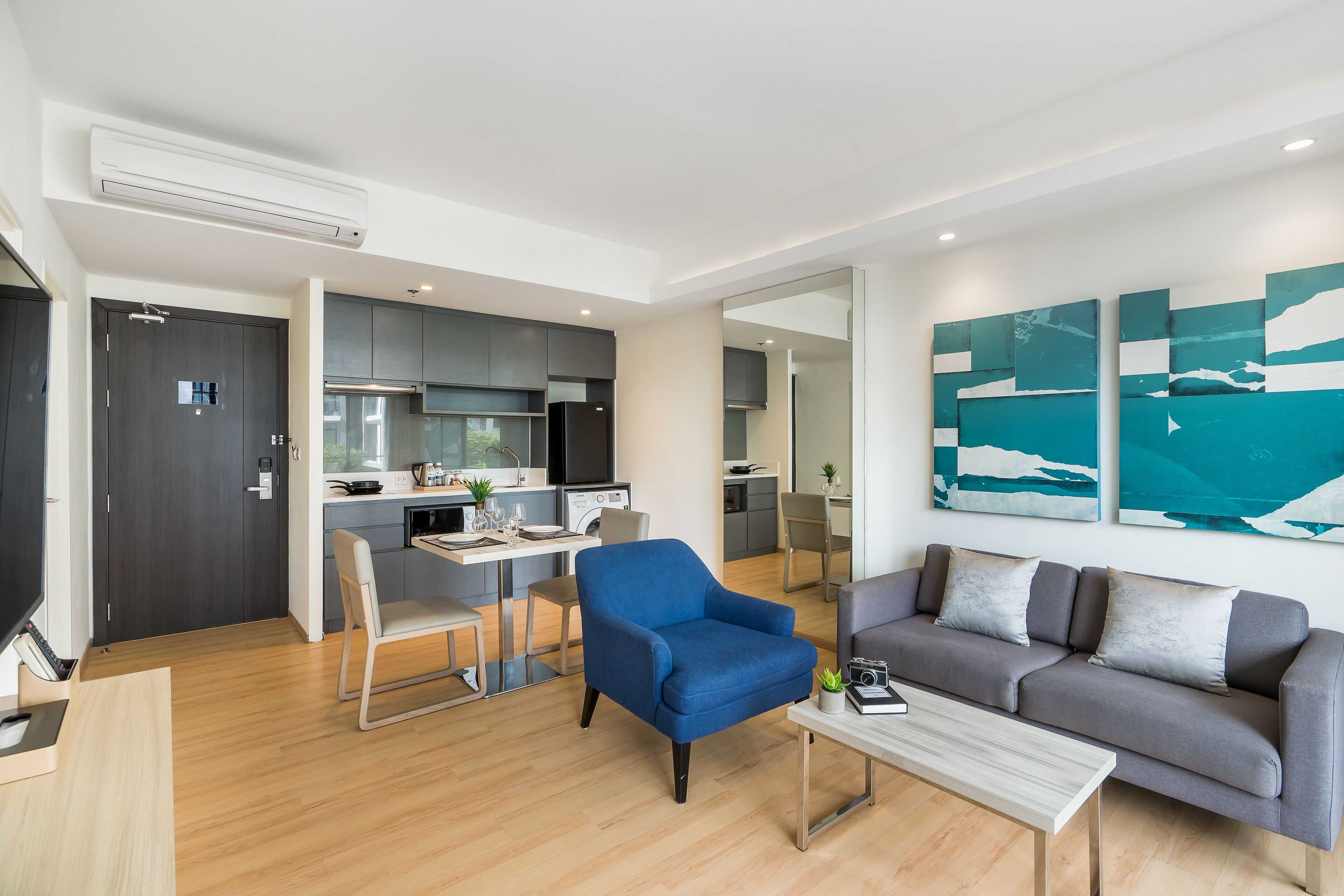 A modern living room with a blue armchair, grey sofa, and a coffee table. There is a kitchen area with grey cabinets and a dining table.