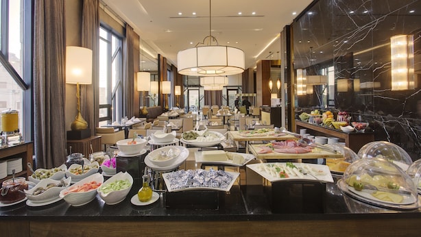 Un comedor de hotel con varias mesas dispuestas para el desayuno, mostrando una variedad de alimentos y una moderna lámpara de araña.