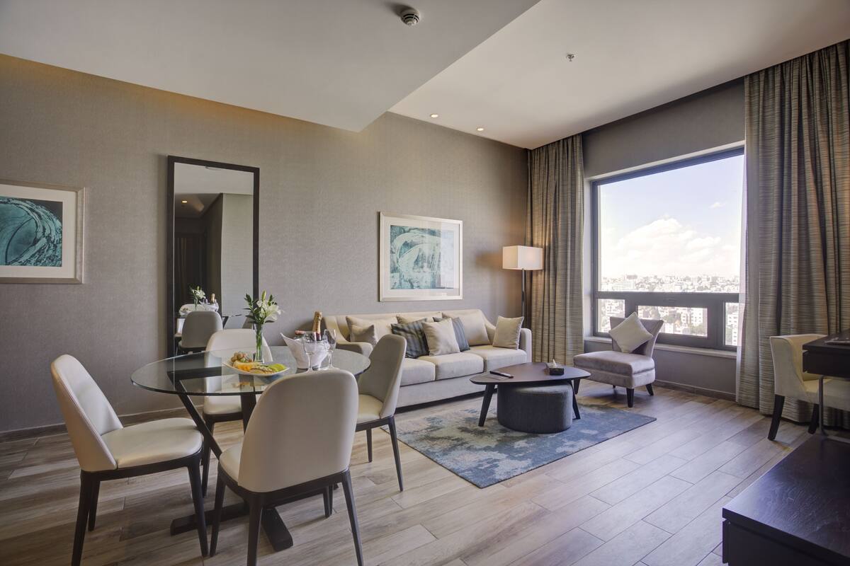 Una sala de estar moderna con una mesa de comedor de vidrio, un sofá y una mesa de centro. Hay dos sillones, una lámpara y obras de arte en las paredes.