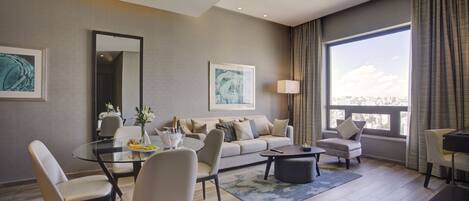 A modern living room with a glass dining table, a sofa, and a coffee table. There are two armchairs, a lamp, and artwork on the walls.