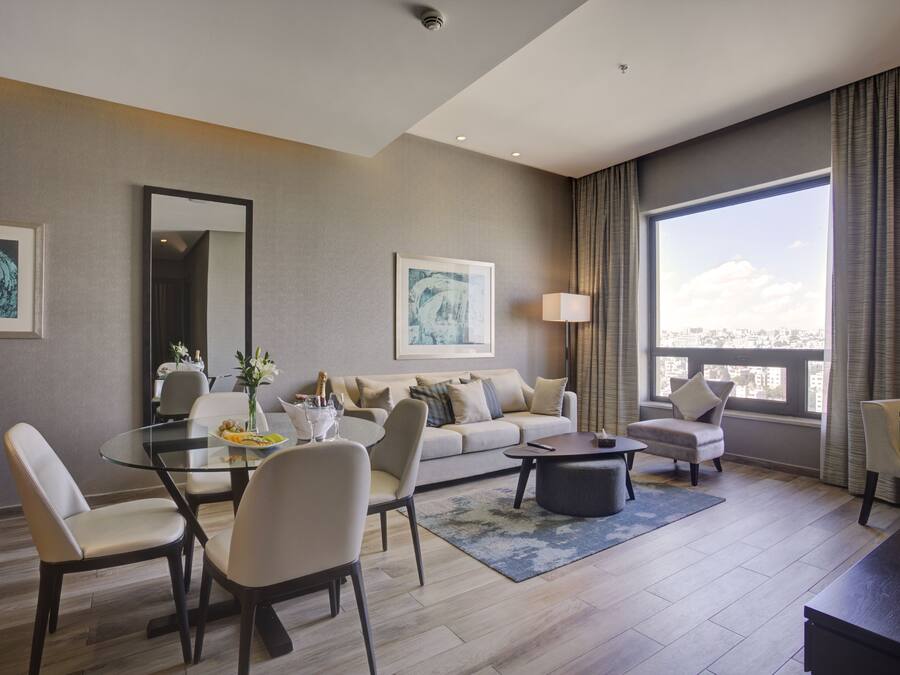 A modern living room with a glass dining table, a sofa, and a coffee table. There are two armchairs, a lamp, and artwork on the walls.