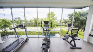 A gym with a treadmill, elliptical, and stationary bike, large windows with a view of greenery and buildings. - Maximum Land & House with WiFi (Udon Thani)