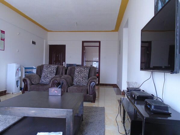 Una sala de estar con un conjunto de sofá, una mesa de centro, un televisor de pantalla plana y una pequeña mesa con dispositivos electrónicos.