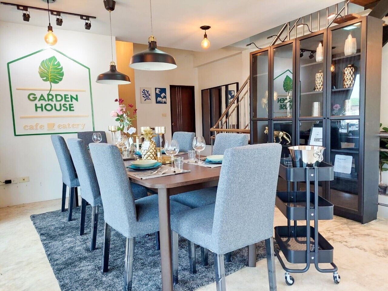 Zona pranzo con tavolo imbandito per sei persone, sedie grigie rivestite in tessuto, illuminazione a sospensione e una vetrina con oggetti decorativi.