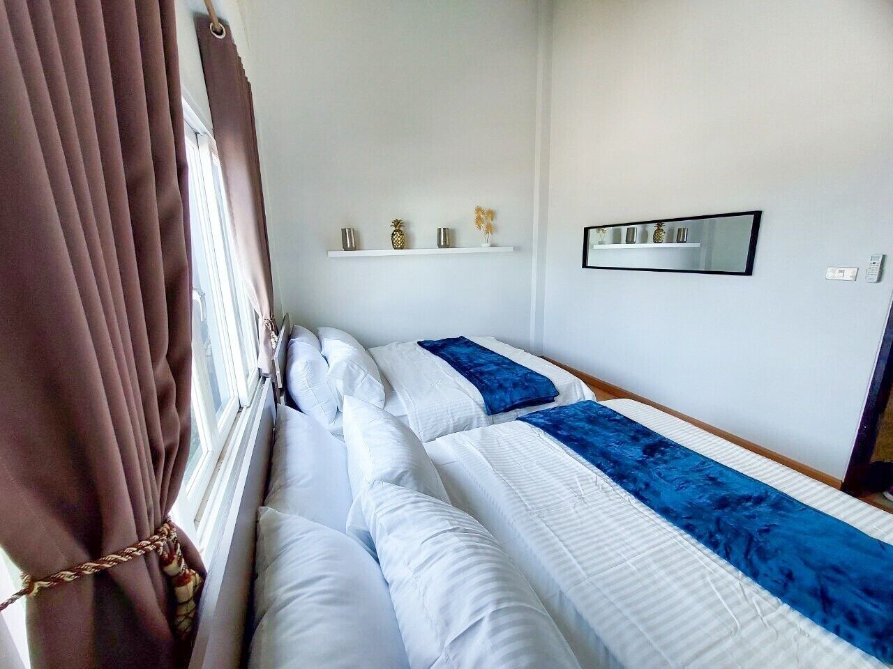 A hotel room with two beds, a window with curtains, a shelf with decorative items, and a mirror on the wall.