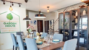 Un comedor con una mesa de madera dispuesta para seis personas, rodeada por seis sillas grises tapizadas. Hay lámparas colgantes sobre la mesa y una escalera con barandilla es visible al fondo.