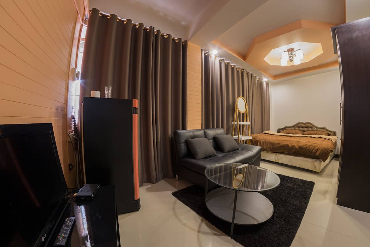 Una habitación de hotel moderna con un televisor de pantalla plana, un sofá, una mesa de centro redonda, una cama con cabecera marrón y un ventanal con cortinas.