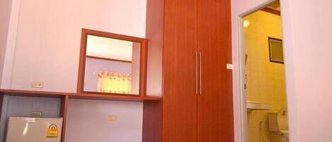 A room with a wooden wardrobe, a small refrigerator, a window with curtains, and a door leading to a bathroom.