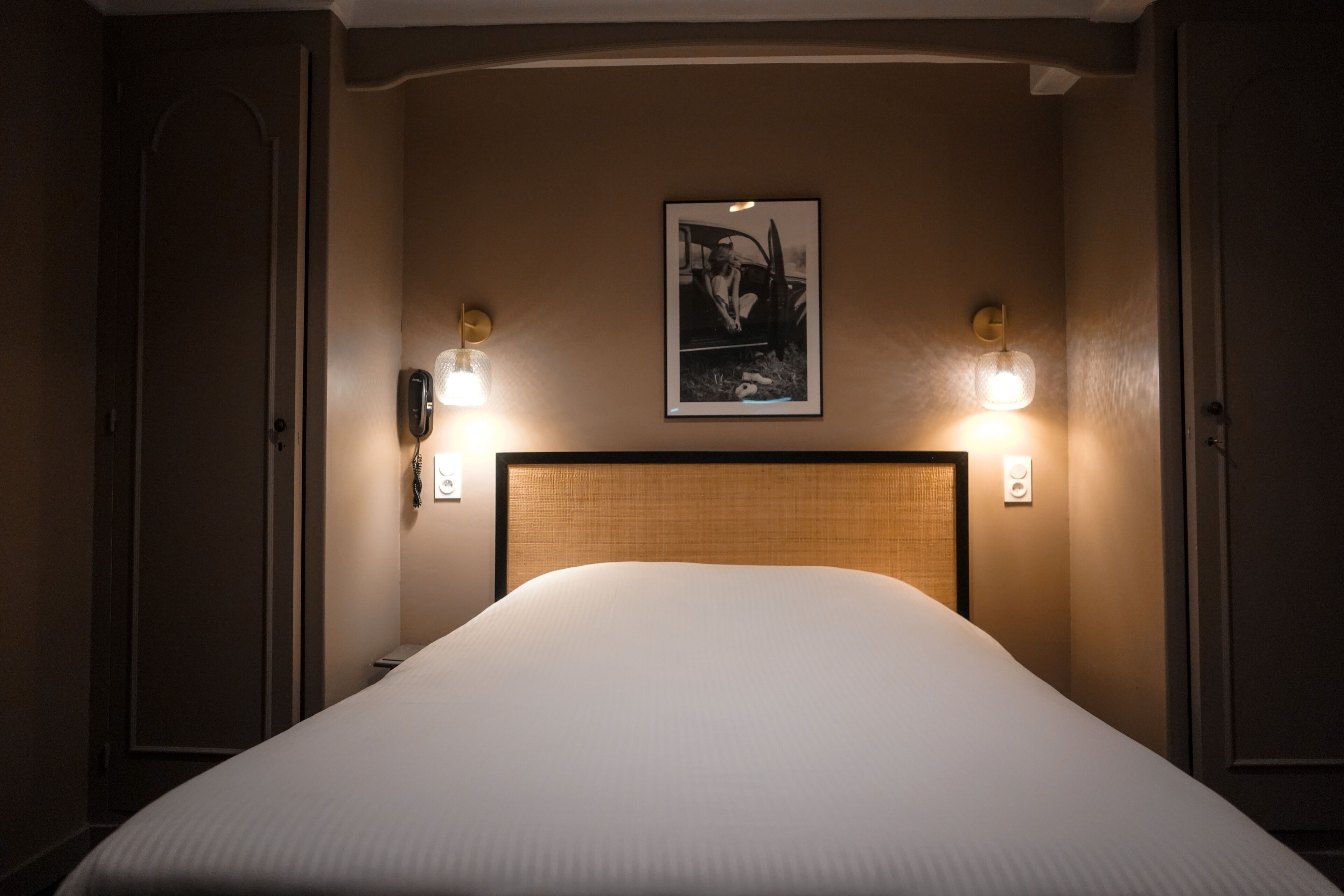 Una cama bien tendida en una habitación de hotel, con un cabecero, dos lámparas de pared y un cuadro enmarcado sobre la cama.