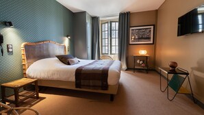 A neatly arranged bedroom with a large bed, a bedside table with a lamp, a small table with flowers, and a television on the wall. - Hôtel Saint Julien (Angers)