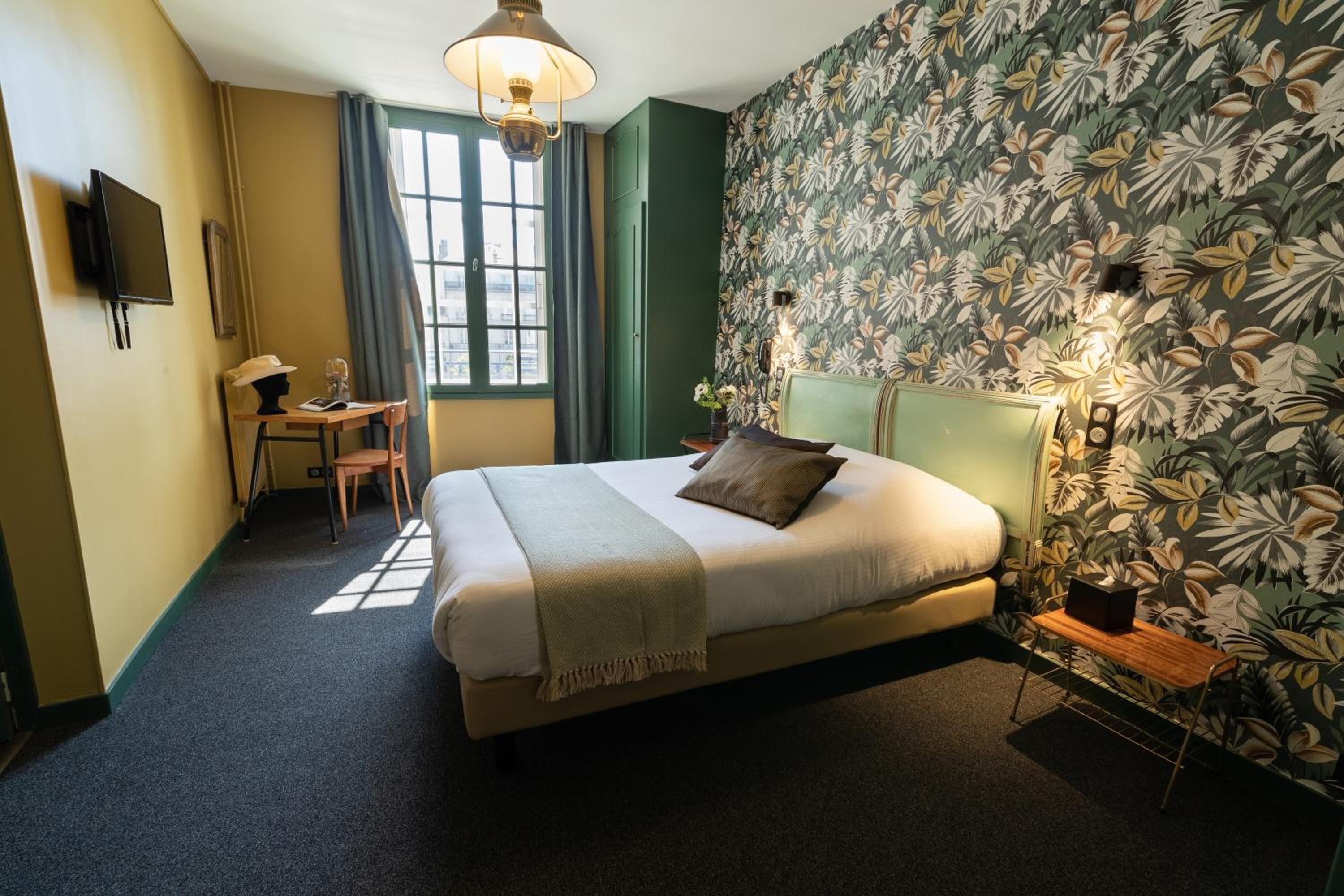 Un dormitorio con cama, escritorio y silla. Hay una ventana con cortinas, una pared con diseño de hojas y una lámpara en el techo.