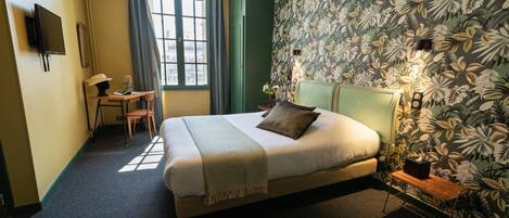 Un dormitorio con cama, escritorio y silla. Hay una ventana con cortinas, una pared con diseño de hojas y una lámpara en el techo.