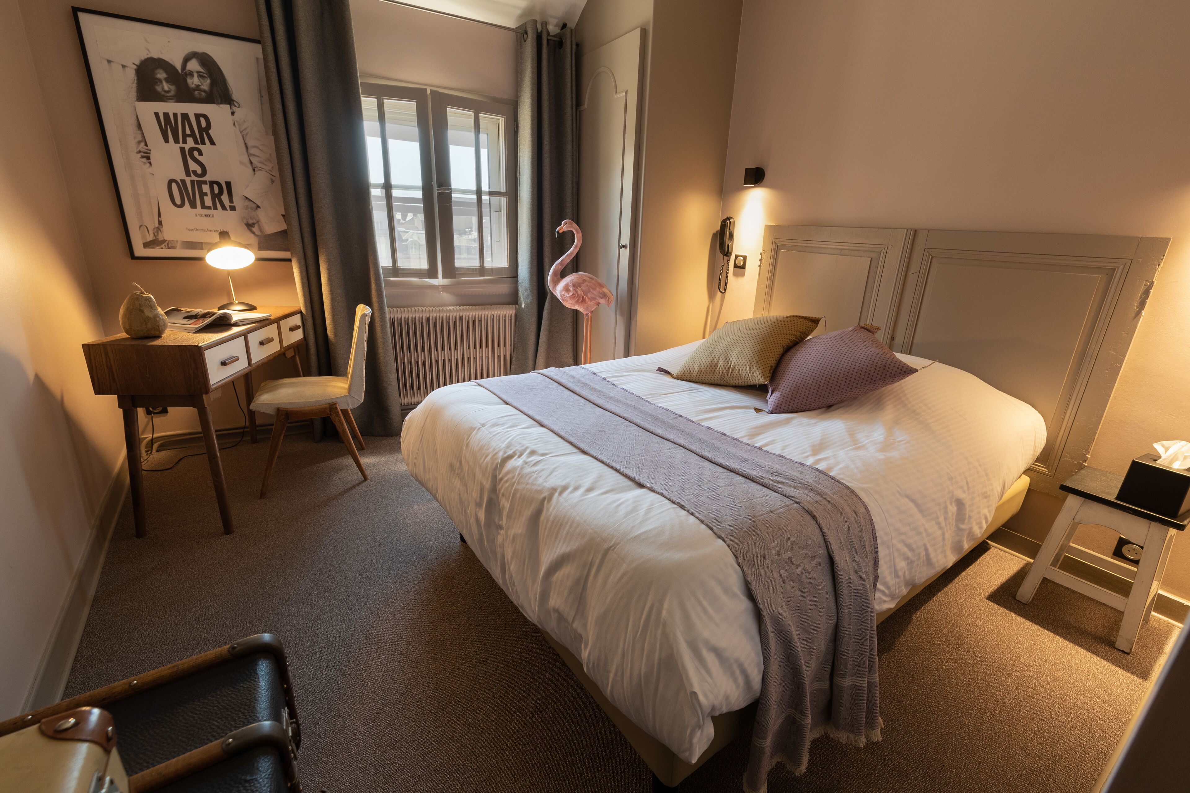 A neatly arranged bedroom with a bed, a desk, a chair, a standing lamp, and a wall-mounted phone.