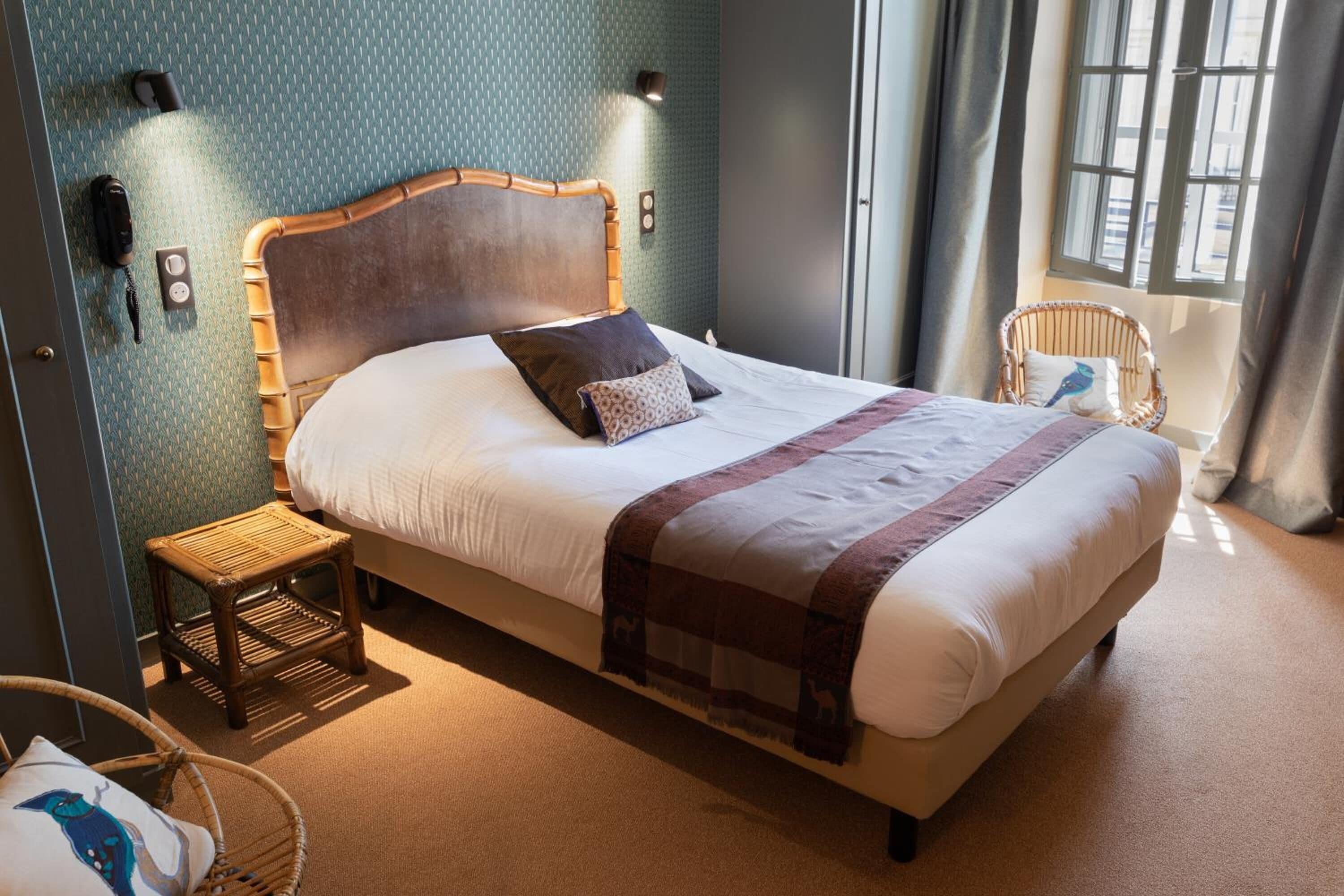 Una cama bien hecha con cabecera de madera, mesita de noche y una silla de mimbre.