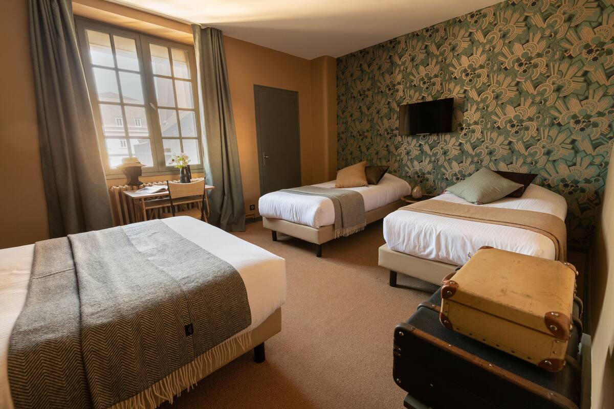 A hotel room with three beds, a desk with a lamp, a television, and a patterned wallpaper.