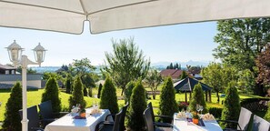 Un espacio exterior para comer con mesas y sillas, rodeado de follaje y vistas a casas y montañas.