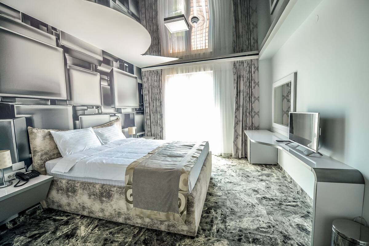 A modern hotel room with a large bed, a TV, a desk, and a large window.