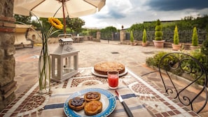 Ein Tisch mit Kuchen, gefüllten Teilchen, einem Glas roter Flüssigkeit und einer Laterne auf einer gefliesten Terrasse mit Blick auf einen Garten und eine Backsteinmauer.