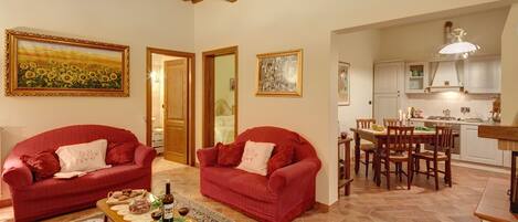 A cozy living room with two red sofas, a dining area with wooden chairs, and a kitchen with white cabinets.