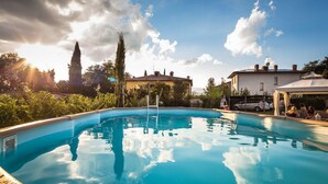 A swimming pool with a clear blue water surface, surrounded by lush greenery and a few buildings in the background. - Cappannelle Country House Tuscany (Castiglion Fibocchi)