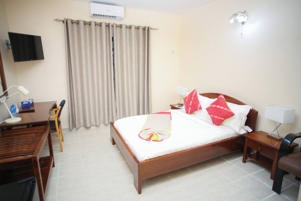 A hotel room with a wooden bed, a desk with a chair, a TV mounted on the wall, and a curtain.