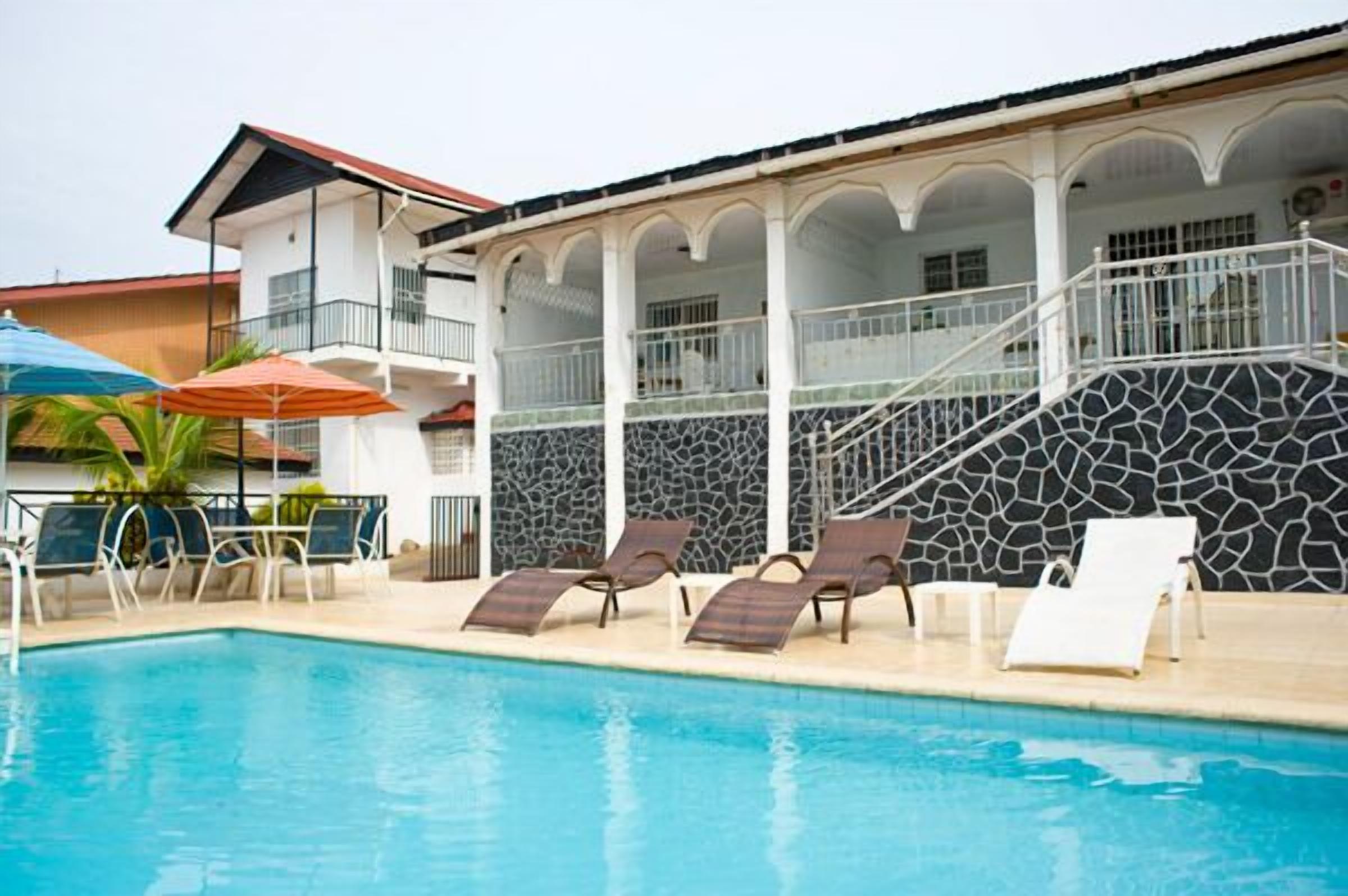 A swimming pool with lounge chairs and umbrellas, a staircase leading to a balcony, and a building with a stone facade.