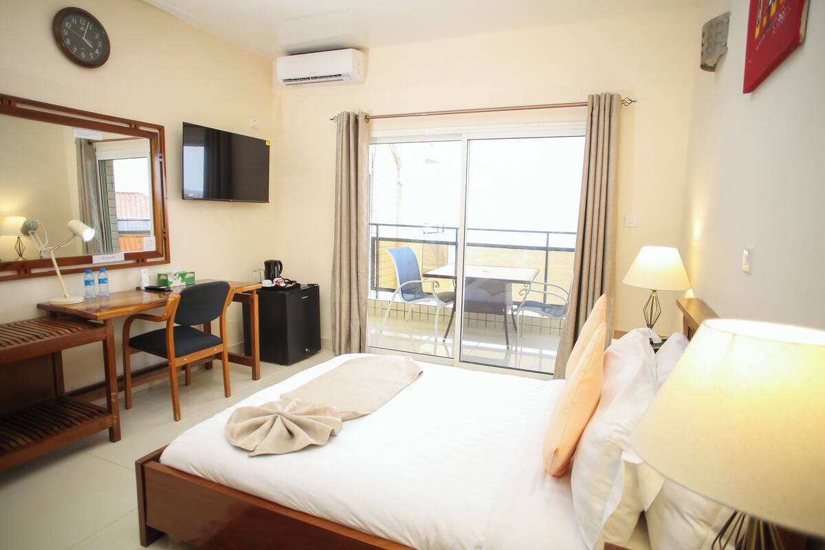 A hotel room with a bed, desk, chair, TV, and a balcony with chairs.