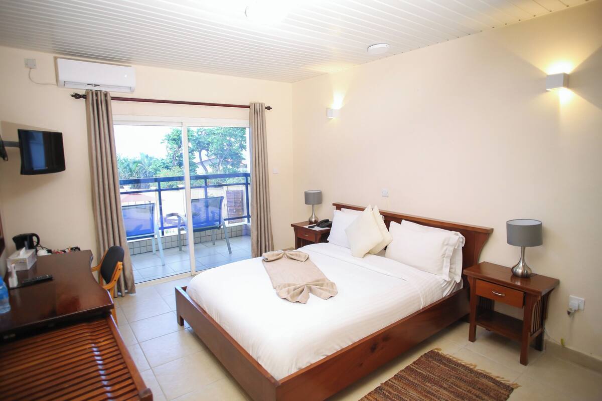 A hotel room with a bed, bedside tables, a desk, a chair, a TV, and a balcony with curtains.