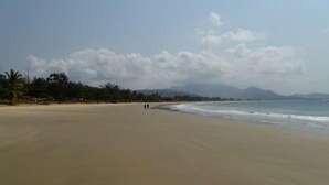 Ein weiter Sandstrand mit einigen Spaziergängern, Palmen und in der Ferne Bergen unter einem teils bewölkten Himmel.