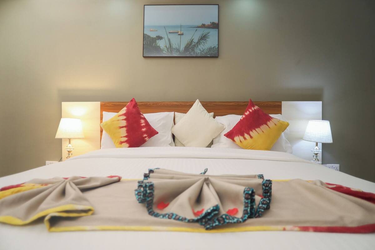 A neatly made bed with a view of the sea and boats, two bedside lamps, and a framed picture on the wall.