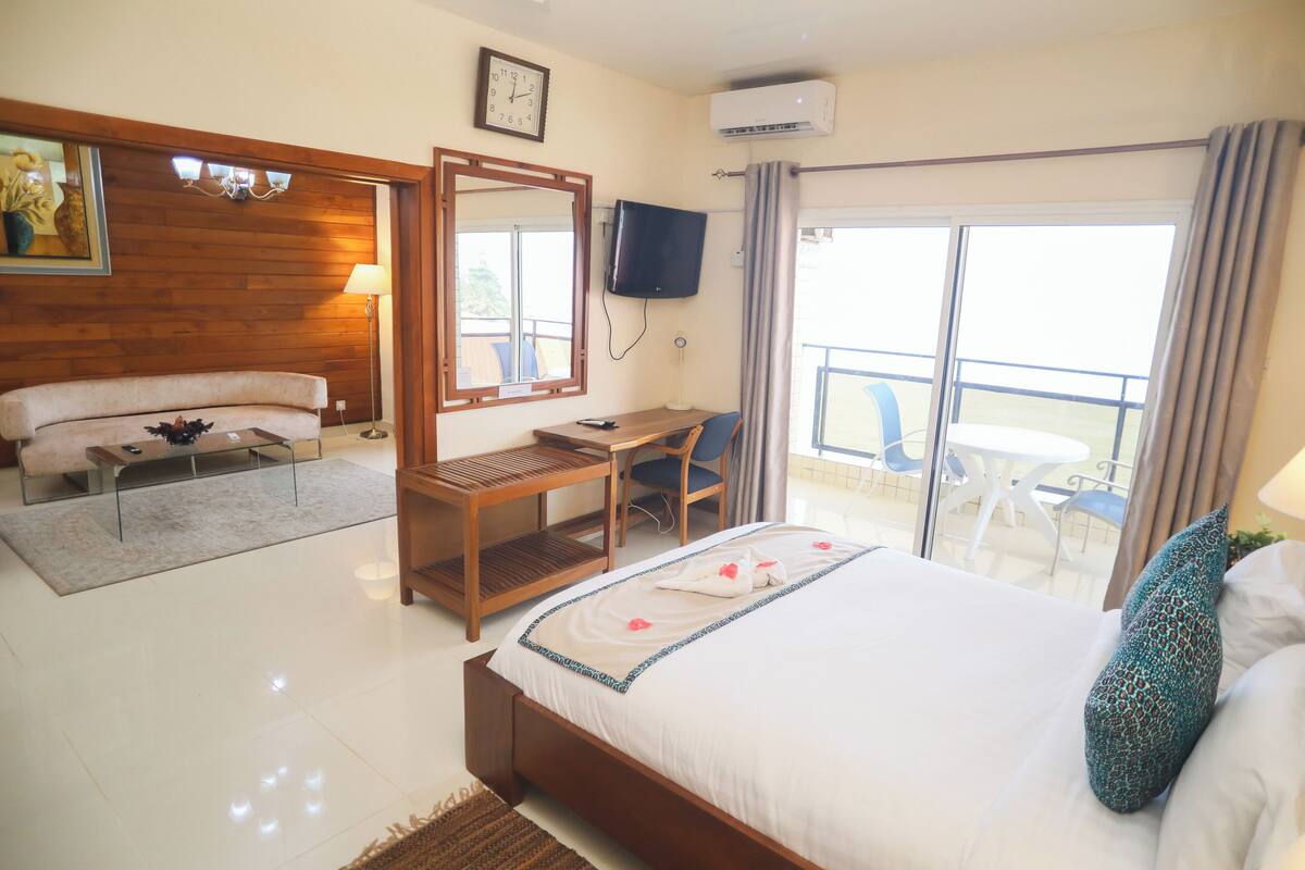 A hotel room with a bed, a desk, a chair, a sofa, a TV, and a balcony with chairs.