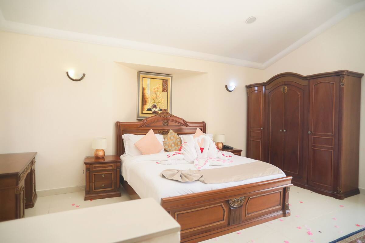 A neatly made bed with pillows and a blanket, a wooden wardrobe, a nightstand with a lamp, and a framed picture on the wall.