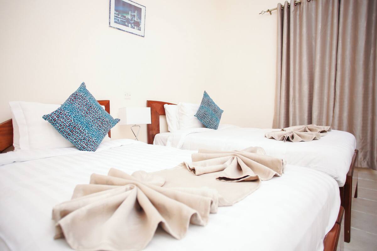A hotel room with two beds, each with a white bedspread and blue patterned pillows. There are beige towels on the beds.