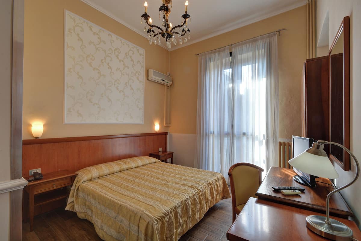 A hotel room with a bed, desk, chair, lamp, air conditioning unit, and a large window with curtains.