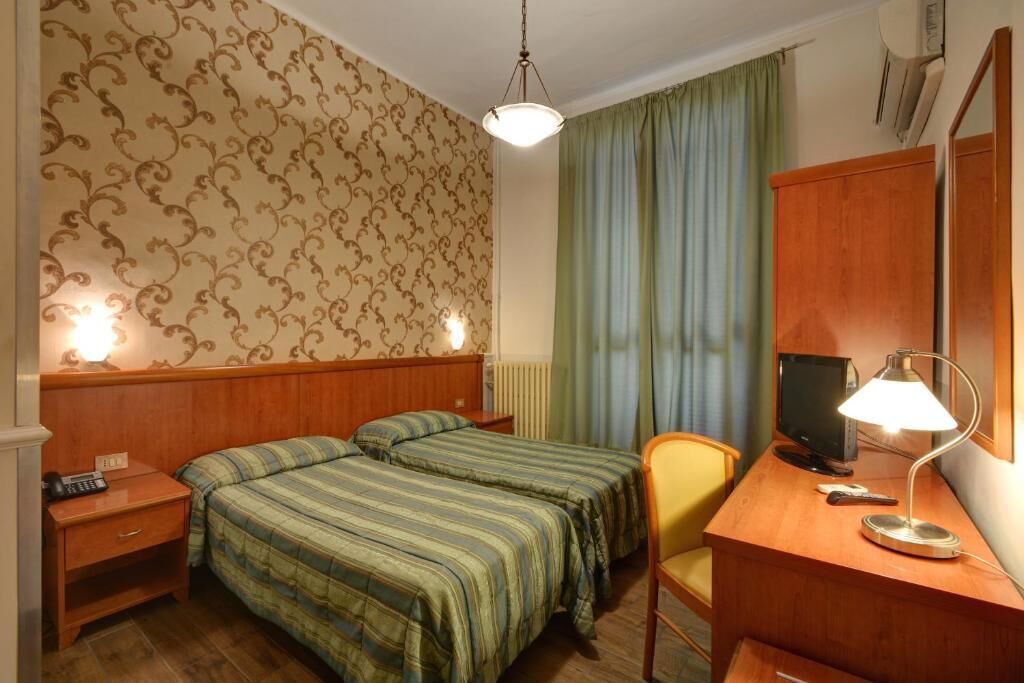 A hotel room with two beds, a desk with a lamp, a chair, a TV, and a window with curtains.