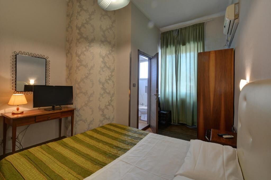 Habitación de hotel con cama, televisor sobre una mesa de madera, un espejo y una puerta que da a un baño.