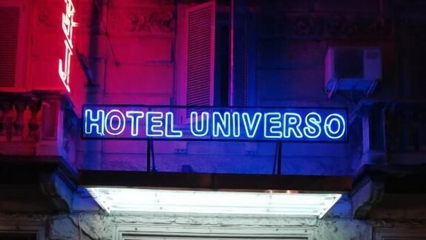 The entrance to Hotel Universo with a neon sign, glass doors, and a visible interior with a reception desk and plants. - HOTEL UNIVERSO (Turin)