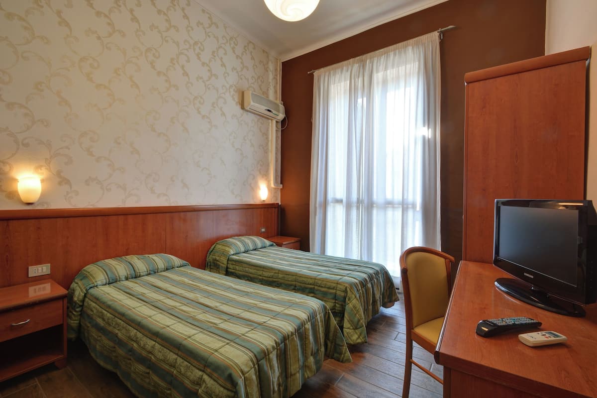 A hotel room with two single beds, a wooden desk with a television and remote controls, a chair, and a window with curtains.