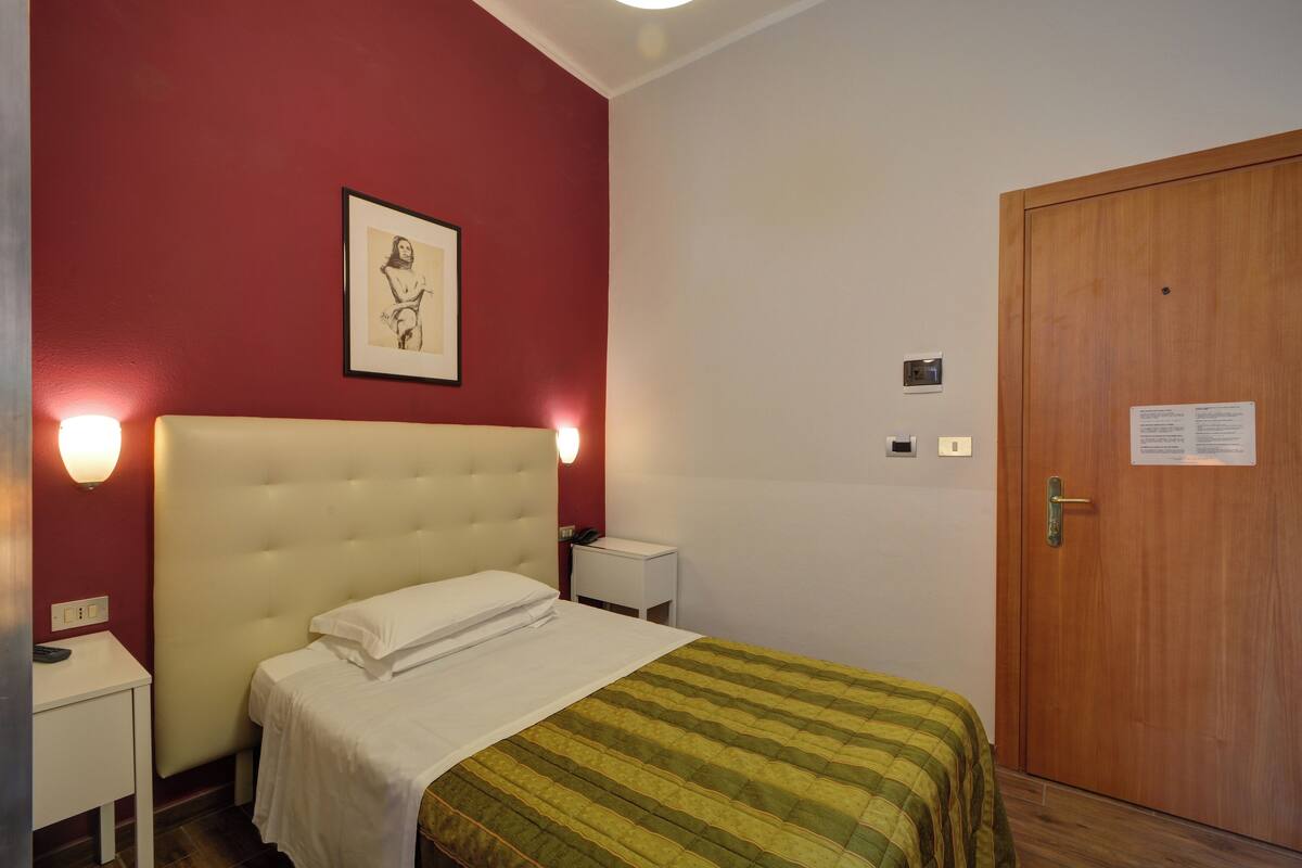 A hotel room with a bed, a bedside table, a nightstand, a wooden door, and a wall-mounted light fixture.