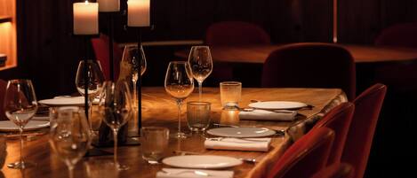A dimly lit restaurant with a long wooden table set for dining, candles, and a red accent lamp.