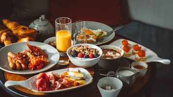 A breakfast table with croissants, a teapot, a glass of orange juice, a bowl of granola with berries, a plate of ham and cheese, and a cup of coffee.
