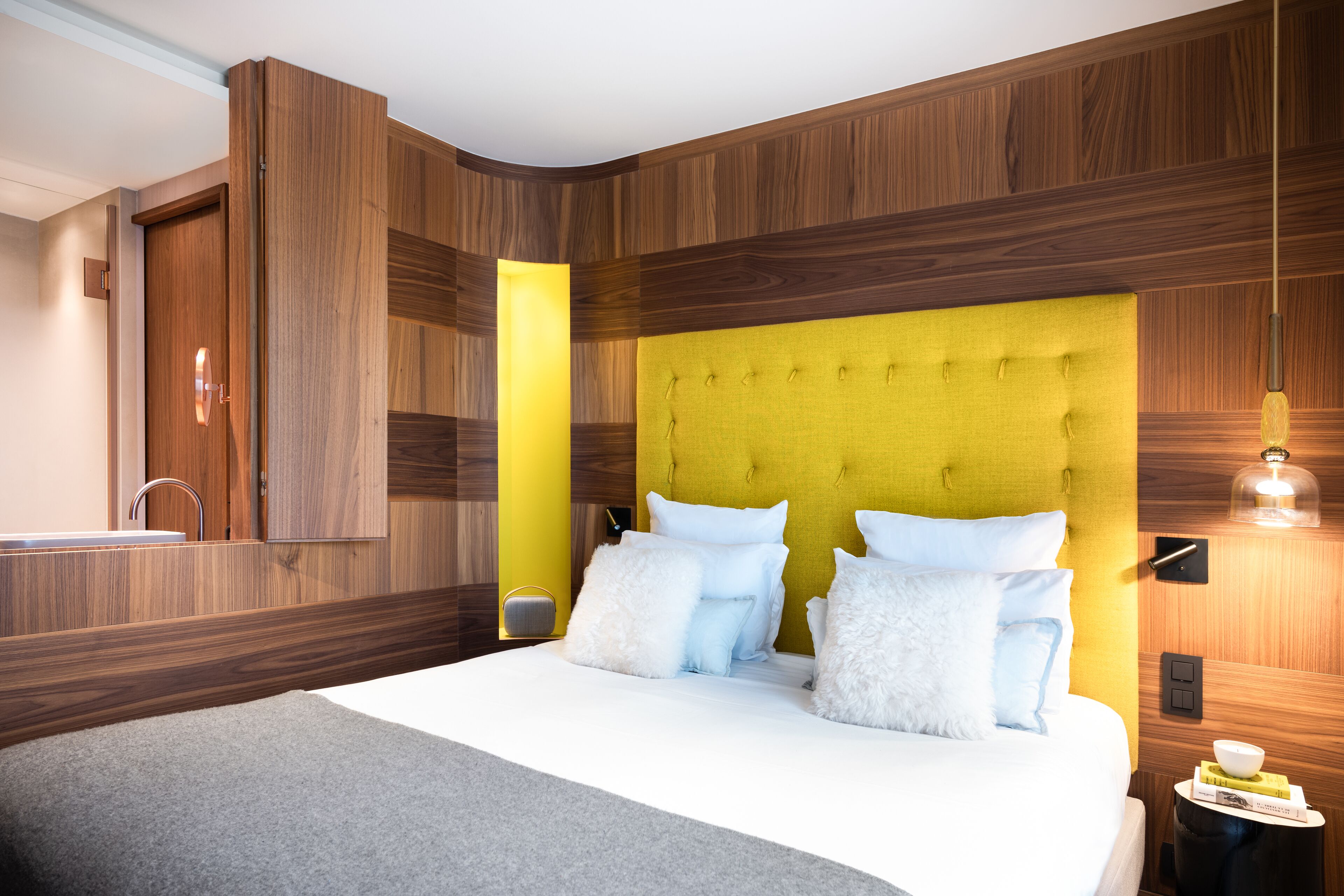 Un dormitorio moderno con cabecera de madera, una mesita de noche con lámpara y otra mesita más pequeña con un libro y una taza.