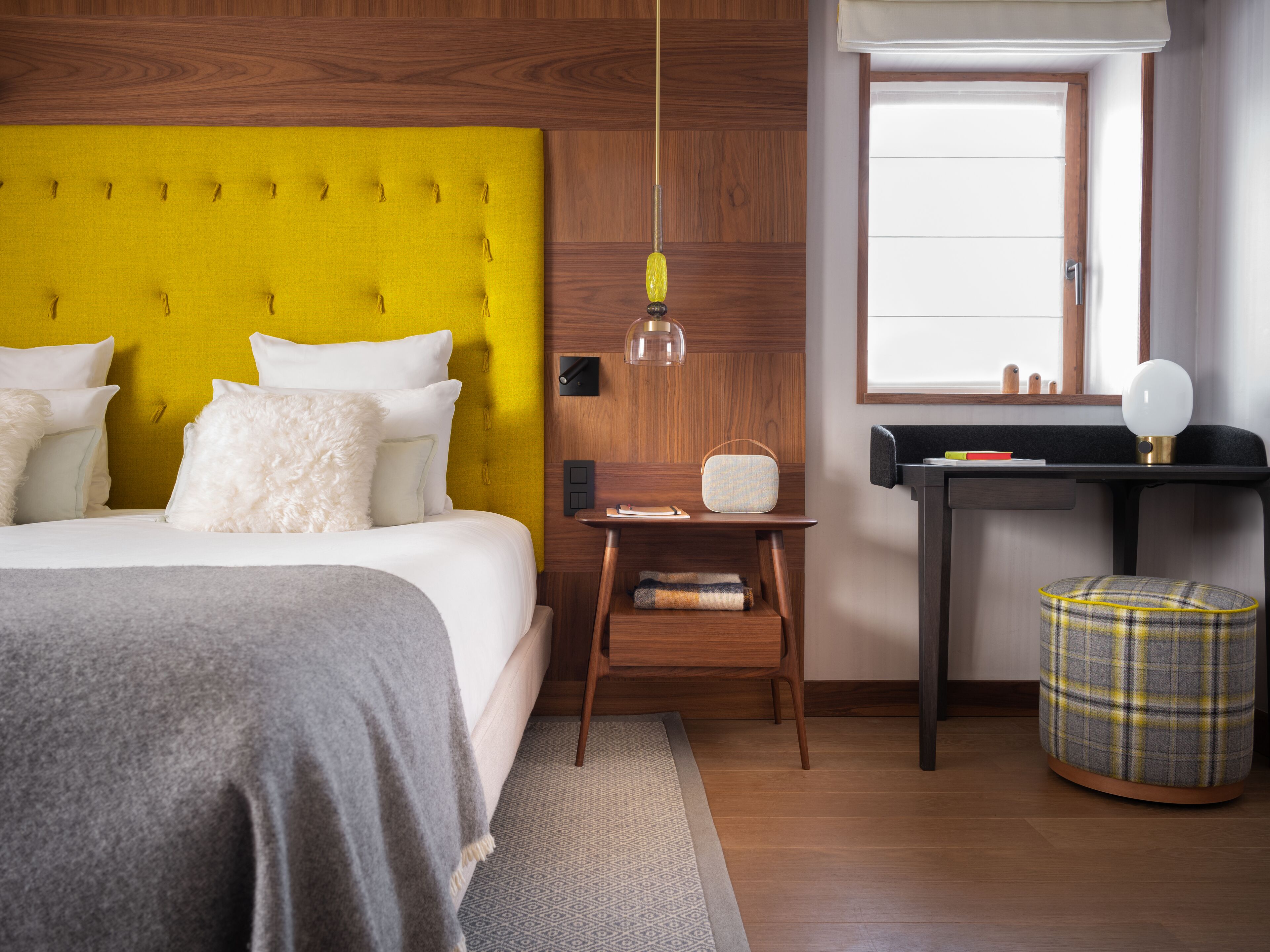 Un dormitorio moderno con un cabecero amarillo acolchado, una mesita de noche de madera, un escritorio con lámpara y un reposapiés escocés.