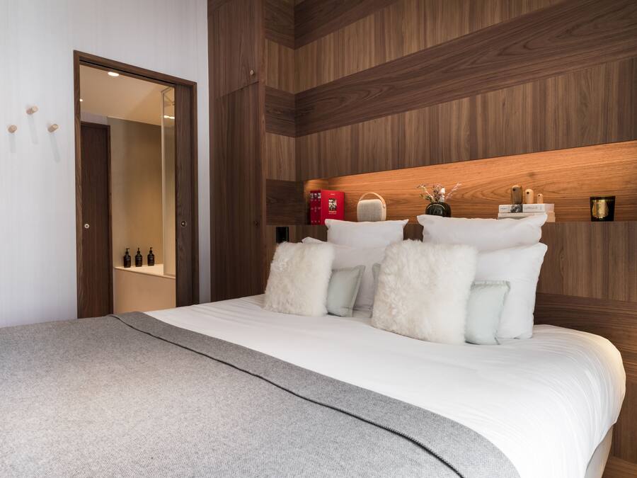 A modern hotel room with a wooden headboard, a large bed with white bedding, a grey throw blanket, and a mirror on the wall.