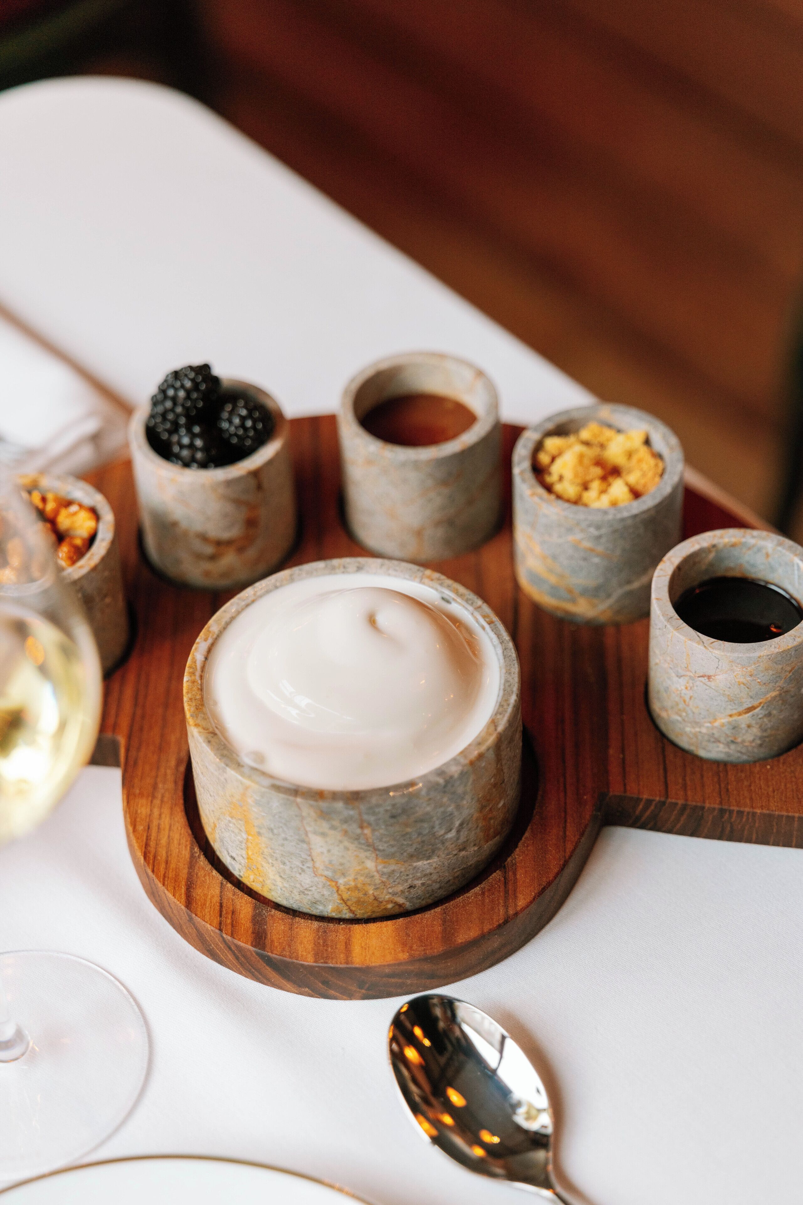 A wooden board with six small containers holding various food items, a glass of white wine, and a spoon on a white tablecloth.