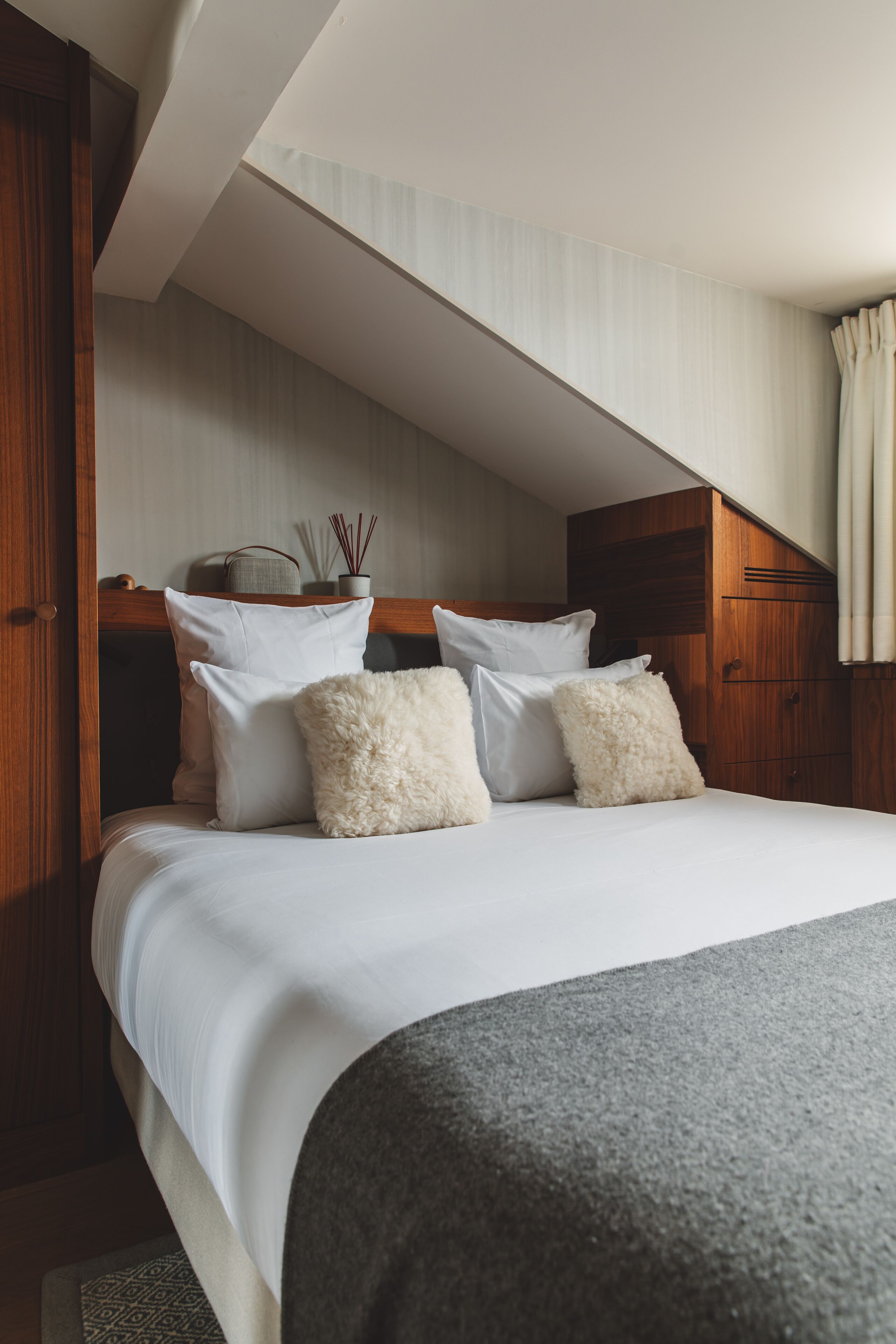 A neatly made bed with white linens and fluffy pillows, a wooden headboard, and a small bedside table with a candle and reed diffuser.