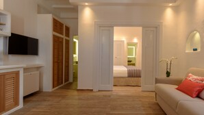 Une chambre d’hôtel moderne avec un plancher en bois, un canapé beige agrémenté de coussins rouges et blancs, un téléviseur à écran plat fixé au mur et une armoire en bois dotée de portes à persiennes.