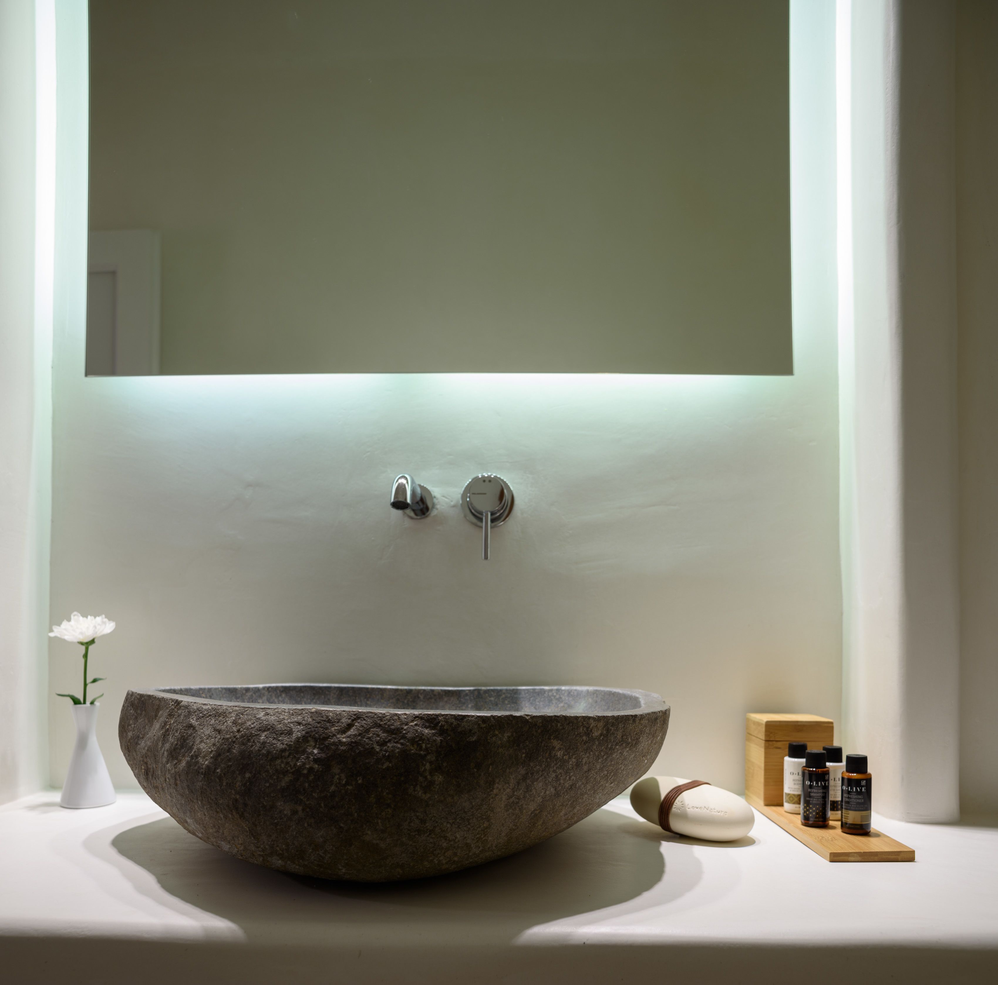 Een moderne badkamer met een grote, ronde stenen wasbak, een spiegel erboven en een klein houten doosje met flesjes ernaast.